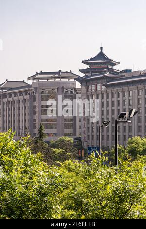 Xian, Cina - 30 aprile 2010: Edifici alti in pietra grigia con accenti di architettura cinese sotto il cielo d'argento dietro il verde fogliame. Foto Stock