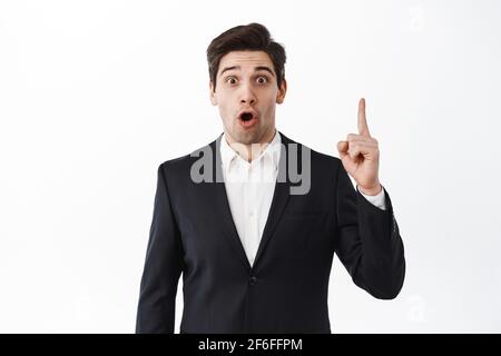 Uomo d'affari sorpreso che punta il dito in su e che gastping stupito, dica WOW, stare eccitato alla macchina fotografica, in piedi in vestito nero contro sfondo bianco dello studio Foto Stock