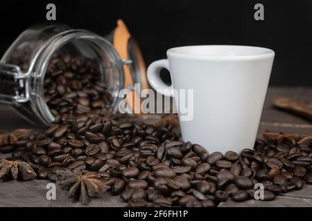 Chicchi di caffè in vaso di vetro con una tazza di caffè bianca. Concetto di caffeina energetica bevanda su sfondo scuro. Foto Stock