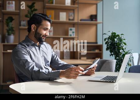 Sorridente uomo d'affari indiano che lavora sul documento di controllo del portatile in ufficio domestico. Foto Stock