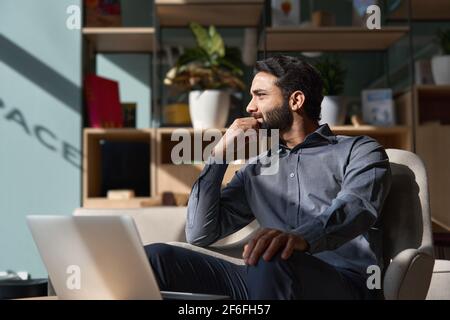 Dirigente indiano uomo d'affari che guarda lontano seduto sulla sedia in un ufficio moderno. Foto Stock