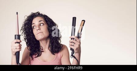 Giovane donna sorridente con capelli ricci non può decidere se usare il suo ferro arricciacapelli o la sua piastra per capelli. Dubbio gesto. Cura e bellezza concept.Young SM Foto Stock