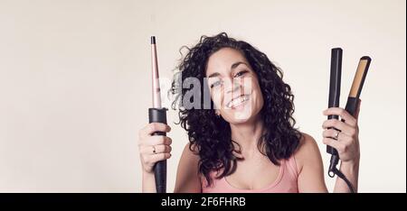 Un giovane sorridente dai capelli ricci non può decidere se utilizzare il ferro arricciacapelli o la piastra per capelli. Spazio copia concetto di cura e bellezza. Foto Stock