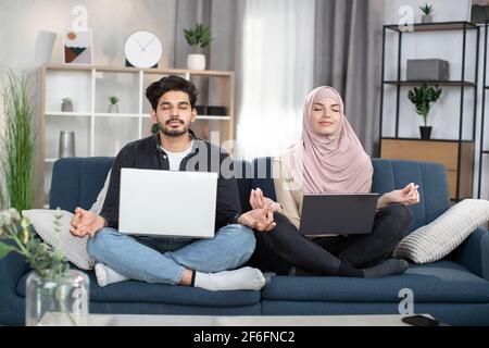 Simpatico giovane musulmano arabo uomo e donna in hijab, seduta in lotus posa con il PC portatile sul divano in soggiorno, e meditando con gli occhi chiusi, dita pieghevoli in mudra. Foto Stock