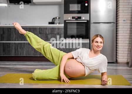 ritratto di bionda caucasica donna incinta sollevamento gamba, facendo esercizi sul tappetino fitness, stretching corpo, impegnato in yoga e sport, mamma-to-be stre Foto Stock