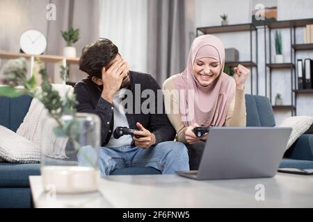 Eccitata divertente coppia musulmana multi ehnic divertirsi insieme mentre si gioca a videogiochi a casa. Ridendo donna in hijab che celebra la vittoria, mentre il suo amico è triste a causa di perso. Foto Stock