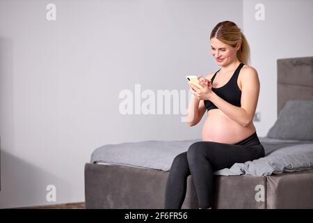 Vista laterale su bella donna incinta seduta con telefono cellulare, avendo riposo da sola, in camera da letto, indossando sport neri, rilassarsi dopo yoga o fitne Foto Stock