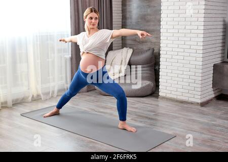 Donna incinta flessibile in leggins e t-shirt allungare le gambe mentre si fa esercizi di yoga durante l'allenamento a casa, stretching corpo Foto Stock