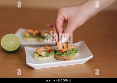 Le donne mettono i gamberi sul bruschetta toast con avocado. Sano antipasto di bruschetta con gamberi guacamole. Lime su un tavolo Foto Stock