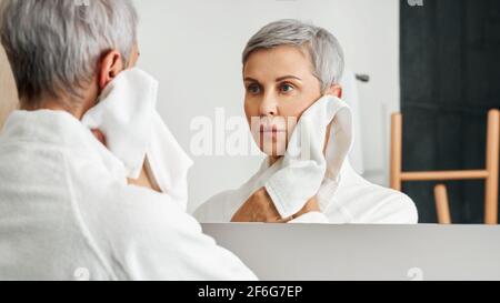 Donna matura con capelli grigi corti guardando il suo riflesso nello specchio del bagno Foto Stock