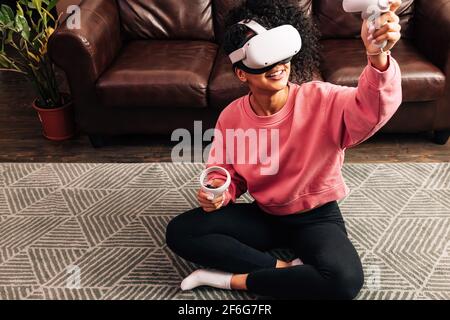 Donna sorridente in salotto con VR set. Giovane donna in abiti comodi che gioca a videogiochi mentre si siede Foto Stock