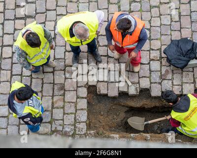 Supervisione importante dei lavori di strada: Un lavoratore scava un buco mentre un altro tiene un pick mentre parla con un supervisore. Altri due guardano sopra. Foto Stock