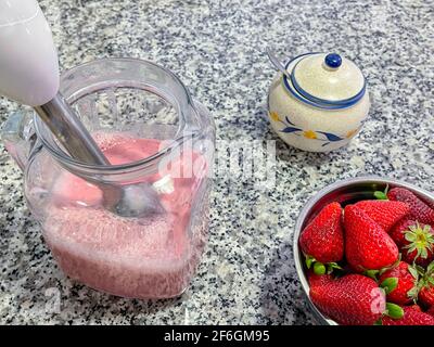 ciotola piena di fragole e una caraffa con frullato di fragola appena fatto, un recipiente per lo zucchero con un cucchiaio all'interno, un frullatore a immersione nel vaso, ingredienti Foto Stock