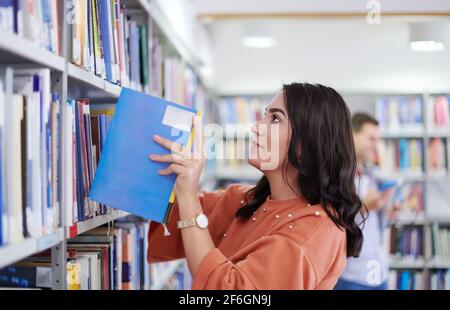 Giovane donna attraente alla ricerca di un libro su una libreria colorata, in una biblioteca. Foto Stock