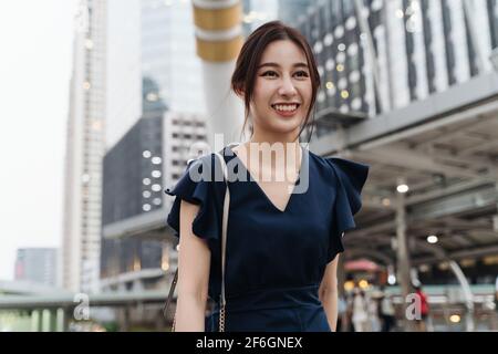 Giovane donna asiatica fiduciosa e indipendente in abbigliamento casual sorridente e camminando lungo la strada della città durante il giorno Foto Stock