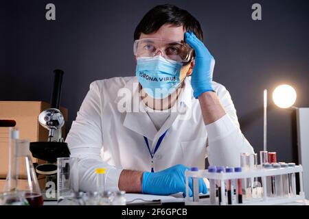 Stanco, esausto, devastato medico scienziato, dopo un lungo turno, testa in mani sulla maschera facciale con il testo coronavirus. Concetto di speranza persa, malattia di Covid-19 Foto Stock