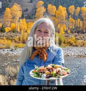 Stati Uniti, Idaho, Sun Valley, ritratto esterno di donna anziana che tiene piatto di insalata mentre si trova lungo il fiume in autunno Foto Stock