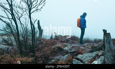 Donna escursionista all'aperto con zaino e abiti spessi nel nebbia su roccia sporgenza aspro natura selvaggia montagne desolate Harz Alpi Norvegia svedese Foto Stock