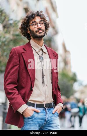 Ritratto di un attraente giovane caucasico in piedi per le strade di Barcellona con le mani in tasca. Ha capelli ricci, barba e occhiali. Abbigliamento casual alla moda Foto Stock