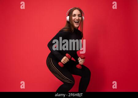 Foto a profilo laterale di bella felice sorridente giovane donna brunet che indossa abiti sportivi neri isolati sulla parete di fondo rossa squating utilizzando manubri Foto Stock