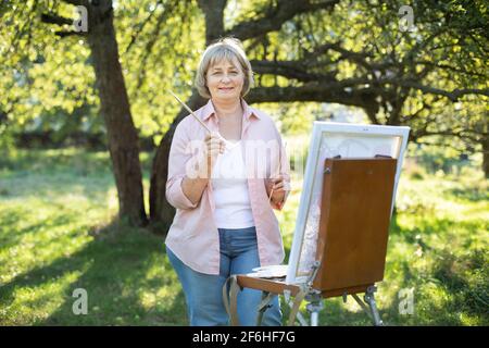 Felice allegro jolly pensò 50-età artista donna, con cavalletto e spazzole, pittura all'aperto in giardino primavera verde la mattina soleggiata. Hobby, ispirazione e concetto di svago. Foto Stock