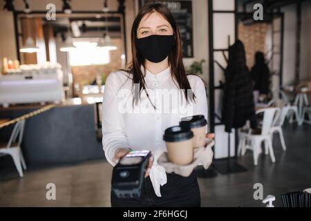 Cassiere in maschera di protezione che tiene la macchina del lettore della carta di credito e due tazze di caffè. Concetto di pagamento senza contatto al caffè Foto Stock