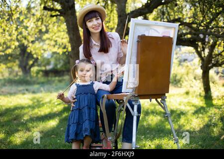 Carino bambina ragazza nel giardino estivo, tiene un pennello e dipinge una foto su tela con la sua mamma insieme. La mamma giovane e graziosa si siede sulla sedia e abbraccia la ragazza, guardando la macchina fotografica con un sorriso. Foto Stock