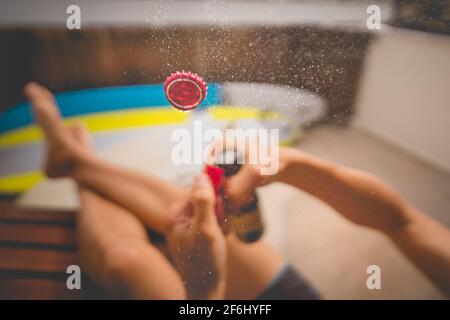 Bere birra fredda e godersi la soleggiata giornata estiva. Bottiglia di birra aperta con spruzzi. Mettere a fuoco il tappo nell'aria. Foto Stock