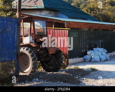 Un vecchio trattore rosso si trova su una strada rocciosa rotta vicino casa privata recintata vecchia Foto Stock