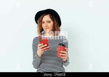 Giovane donna confuso e imbarazzata che indossa un cappello nero e una camicia a righe che tiene una tazza di caffè rossa guardando nel suo smartphone, sfondo bianco studio. Foto Stock