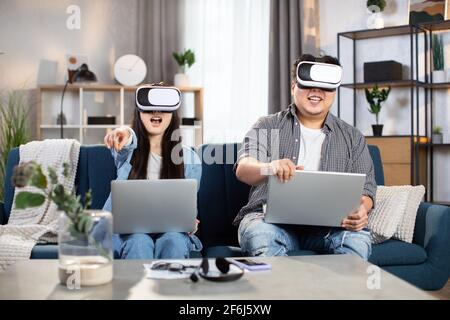 Giovane coppia asiatica in abbigliamento casual che utilizza il visore VR e il laptop per giocare durante il tempo libero a casa. Concetto di famiglia e intrattenimento. Foto Stock