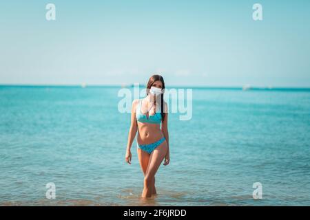 Estate. Una giovane bella donna che indossa una maschera protettiva si pone in piedi in riva all'oceano. Il concetto di vacanze estive durante la pandemia del coronavirus. Foto Stock