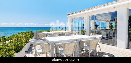 Immagine orizzontale di un ristorante all'aperto vuoto vicino al mare. Sedie in vimini e tavolo in legno all'interno della zona costiera caffè terrazzato durante il giorno caldo e soleggiato. Foto Stock