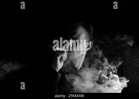 Uomo che fuma sigaro con un bicchiere di whisky nel Mano su sfondo nero scuro isolato.Creative fotografia in bianco e nero ritratto con una nuvola di Foto Stock