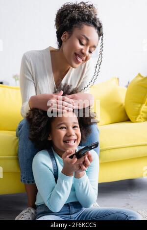 eccitato capretto afroamericano che tiene il telecomando mentre guarda la tv vicino a madre felice Foto Stock