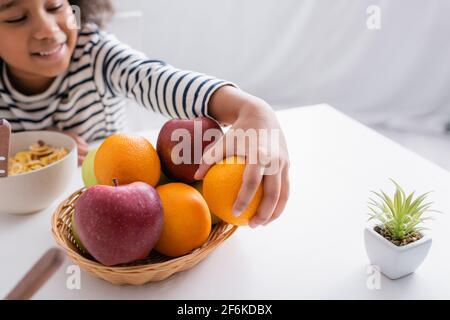 sorridente afroamericana ragazza che prende arancio maturo dal cesto di vimini in cucina, sfondo sfocato Foto Stock