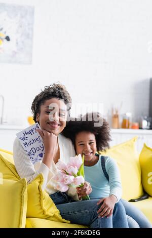 gioiosa ragazza afroamericana seduta vicino a mamma felice che tiene tulipani e buon giorno delle madri card Foto Stock