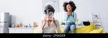 spaventata donna afro-americana che gesturing in vr visore vicino a allegra figlia, banner Foto Stock