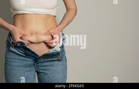 Donna in jeans e camicia bianca che misura il suo grasso del ventre. Donna`s figura closeup studio RAW girato in fondo grigio. Dieta e concetto di perdita di grasso. Foto Stock