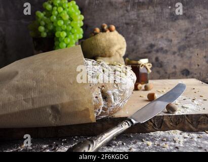 Pane scuro a grana intera in carta da imballaggio. Vista laterale. Uve verdi, noci e miele sullo sfondo. Stile provenzale. Foto Stock