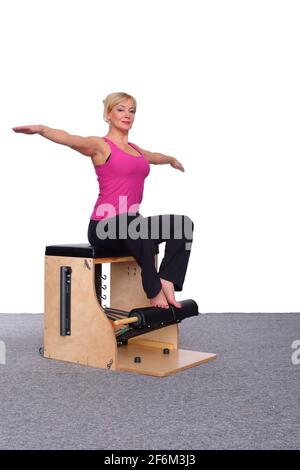 Un addestratore di 50-year-old pratica Pilates su una sedia dell'elevatore, sollevando le sue braccia all'altezza della spalla e raddrizzandosi a suo lato. Seduto Foto Stock