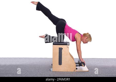 Un addestratore femminile di 50-year-old pratica Pilates su una sedia dell'ascensore, sollevando la sua gamba alta sopra la sua testa. La donna si appoggia sul ginocchio e sulle mani Foto Stock