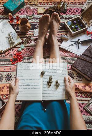 Vista dall'alto di una donna asiatica che tiene un notebook aperto con dadi metallici seduti su un plaid circondato da attributi di gioco di ruolo. Foto Stock