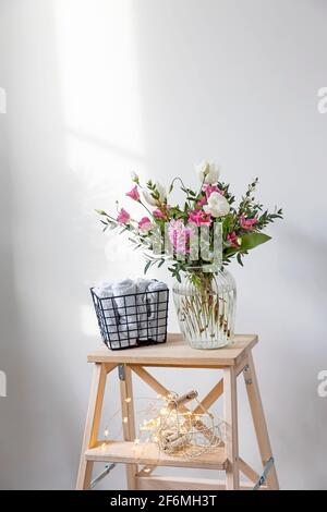 Un bouquet minimalista di tulipani bianchi, eustoma rosa, giacinto, eucalipto in vaso di vetro scanalato su una sedia. Piccoli asciugamani bianchi per le mani e il viso Foto Stock