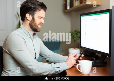 Uomo che prende una videochiamata sul suo cellulare che lavora da casa su un computer desktop. Foto Stock