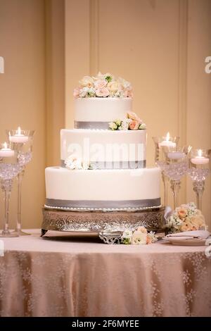 Cerimonia nuziale torta a tre livelli fatta con glassa bianca e. rifiniture grigie decorate con rose bianche e rosa su una tavolo coperto con tovaglia di seta Foto Stock