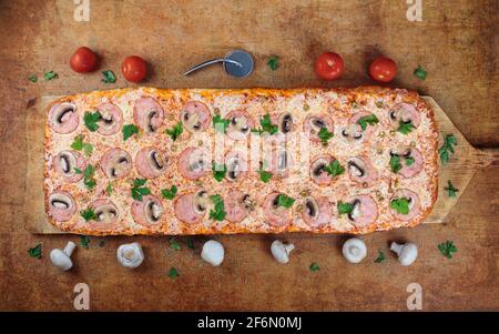 PizzaLunga pizza con prosciutto e funghi si trova su un tavolo di legno. Pizza romana su una spatola di legno. Pizza lunga un metro. Foto Stock