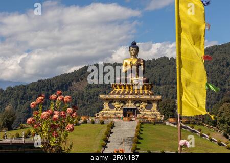 Statua di Buddha con cielo blu e nuvole sullo sfondo e. Fiori accanto a una bandiera di preghiera in primo piano a Ravangla in Sikkim India il 04 novembre 2016 Foto Stock