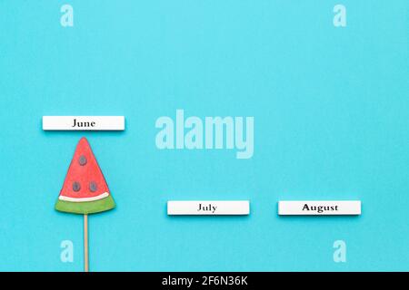 Calendario in legno mesi estivi Giugno, Luglio, Agosto e cocomero lollipop su bastone su sfondo blu. Vacanza concettuale, vacanze o Ciao giugno. Creati Foto Stock