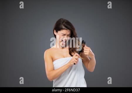 Pettinatura dolorosa dei capelli aggrovigliati con la spazzola per capelli. Donna avvolta in un asciugamano bianco intorno al suo corpo si trova di fronte a una parete grigia e utilizza un pettine per capelli a poppa Foto Stock